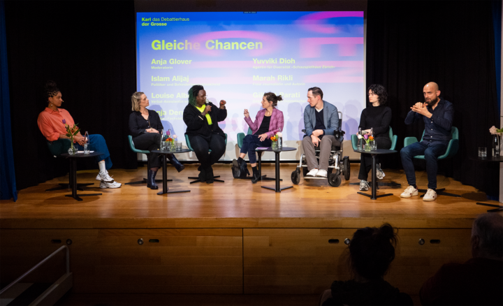 Podiumsdiskussion auf der Bühne im Saal zum Thema "Gleiche Chancen". 7 Personen aus unterschiedlichen Branchen sprechen miteinander. Ein Gebärdensprachdolmetscher übersetzt das Gespräch.